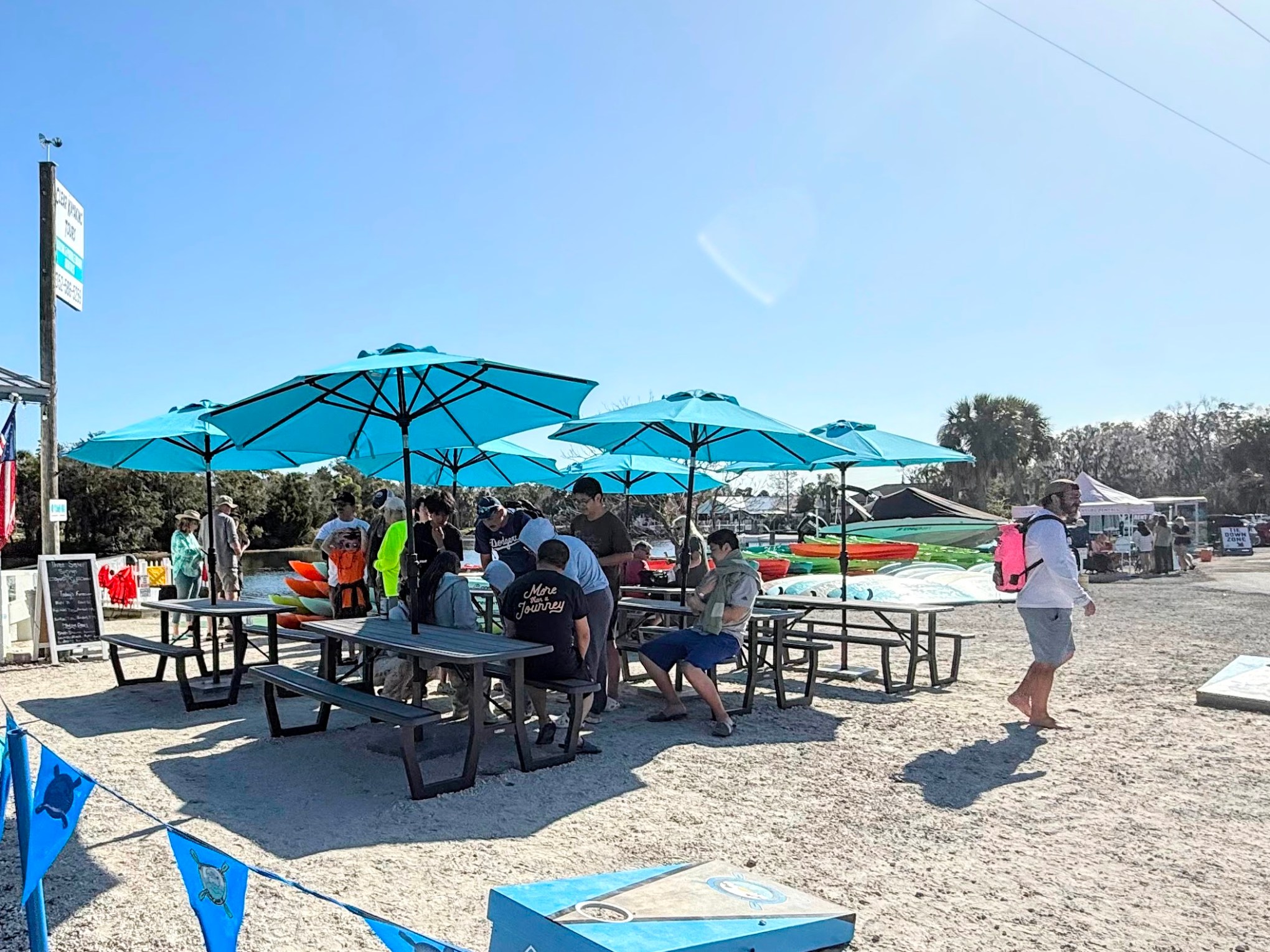 Outdoor shop with blue umbrellas and picnic tables; people are gathered, and kayaks are nearby.