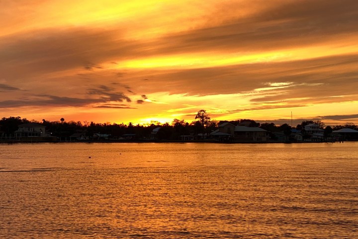 Silhouetted houses against an orange and yellow sunset over a calm lake.
